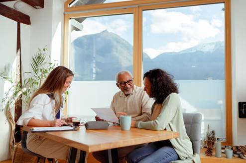 An advisor meets with a mature couple in their bright sunroom. The group sits around a wooden table and reviews